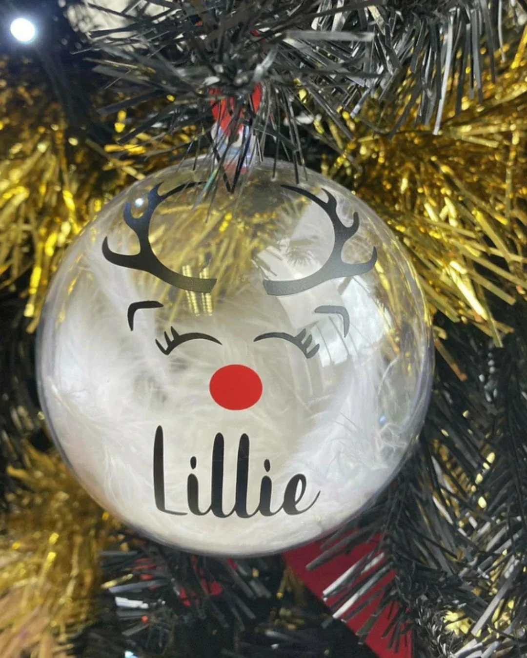 A clear bauble with a reindeer face and the name 'Lillie' hanging on a gold tinsel.