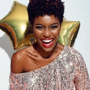 Photo of Laughing Woman with Confetti on Her Hair and Shoulders Posing In Front of White Background While Holding Golden Star Balloons Behind Her Back