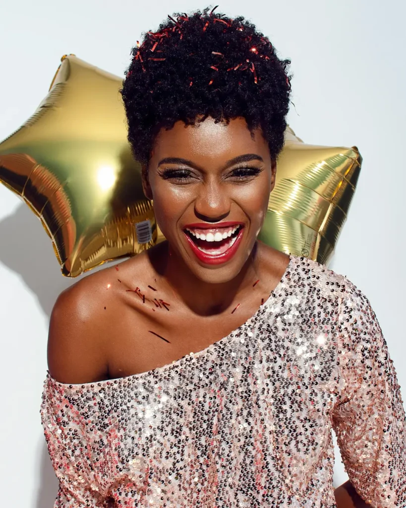 Photo of Laughing Woman with Confetti on Her Hair and Shoulders Posing In Front of White Background While Holding Golden Star Balloons Behind Her Back