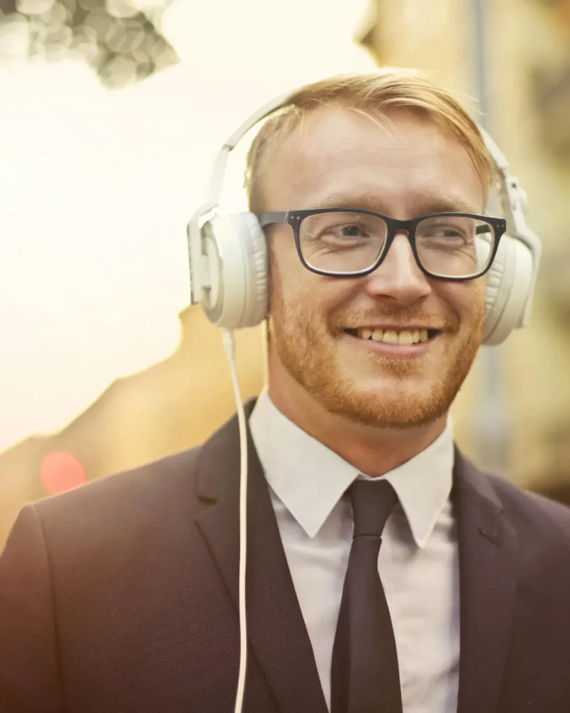 Selective Focus Photo of Smiling Man in a Black Suit and Black Framed Eyeglasses Listening to Music on White Headphones