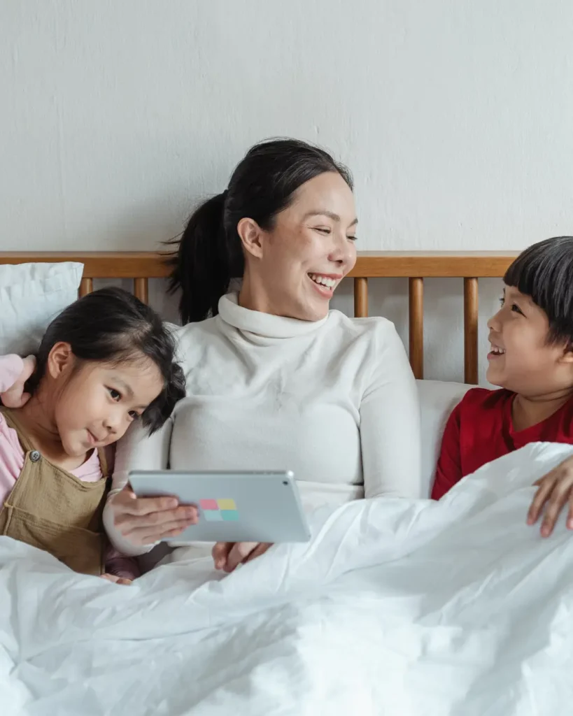 Cheerful Asian mother and little children with tablet having fun in bed