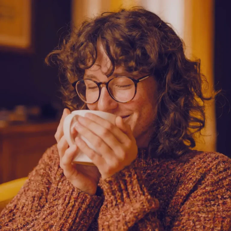 Content young woman with curly hair and glasses smiles with closed eyes while sipping a warm drink from a mug in a cosy armchair